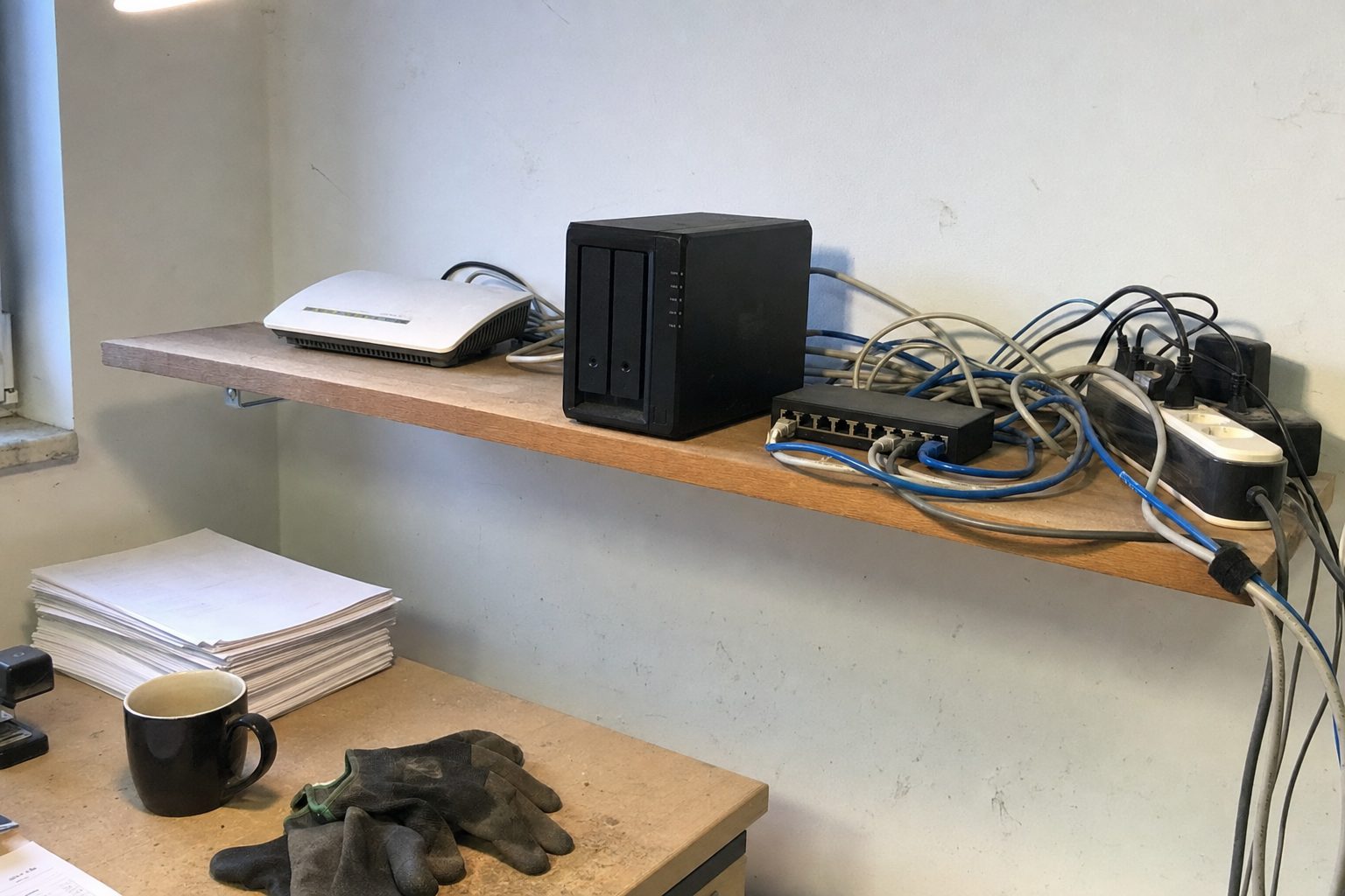 Wandregal im Büro eines Handwerksbetriebs mit Router, NAS-Speicher, Switch und Kabeln, darunter ein Schreibtisch mit Lieferscheinen und Arbeitshandschuhen